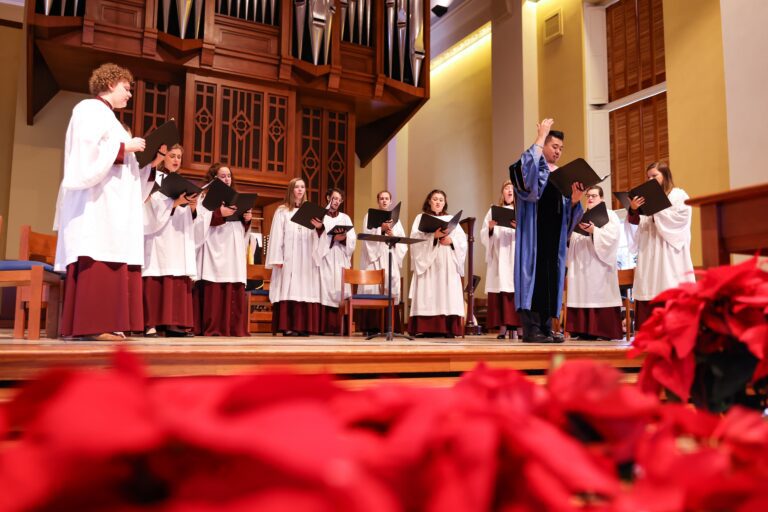 An upward angled photo of Chamber Choir performing during Lessons and Carols, December 2022. The photo is framed on the bottom by red poinsettias. 