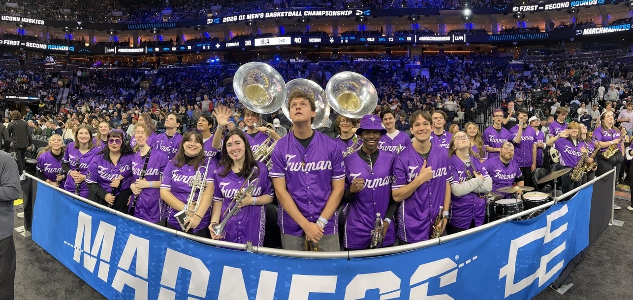 Basketball band standing as a group with their instruments behind the March Madness banner
