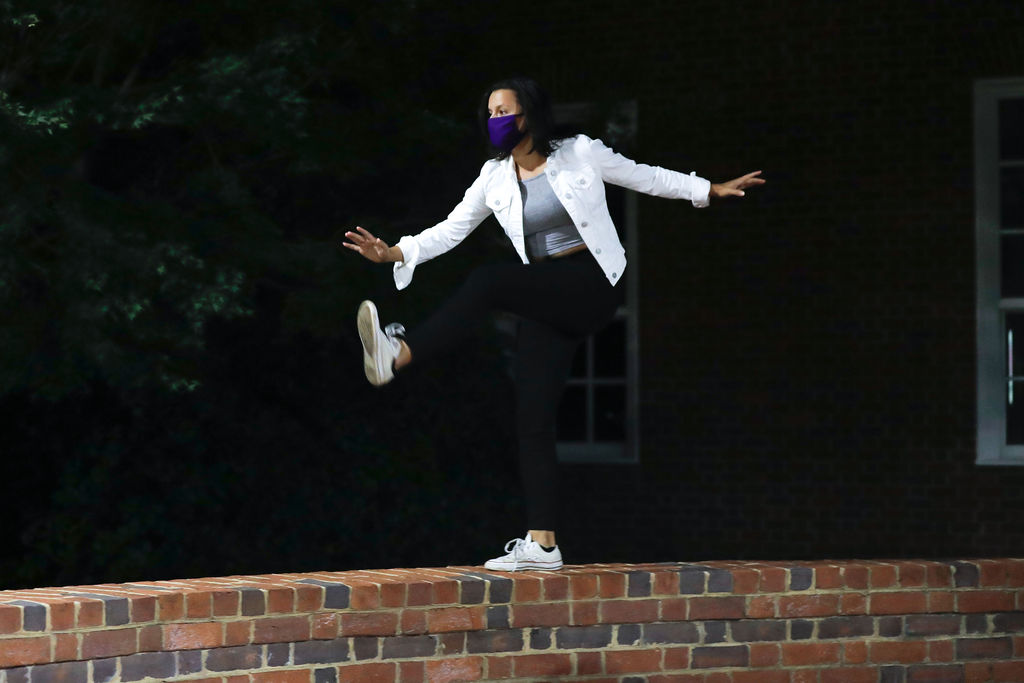 image contains student dancing on a brick wall with white jacket, black pants, and purple mask
