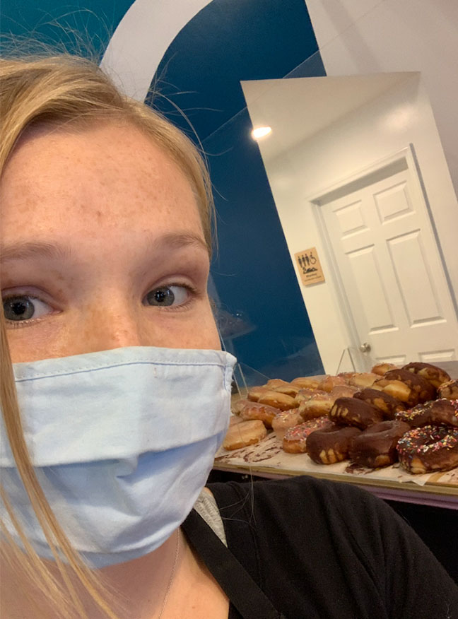 student wearing face mask takes a selfie by a counter top full of donuts