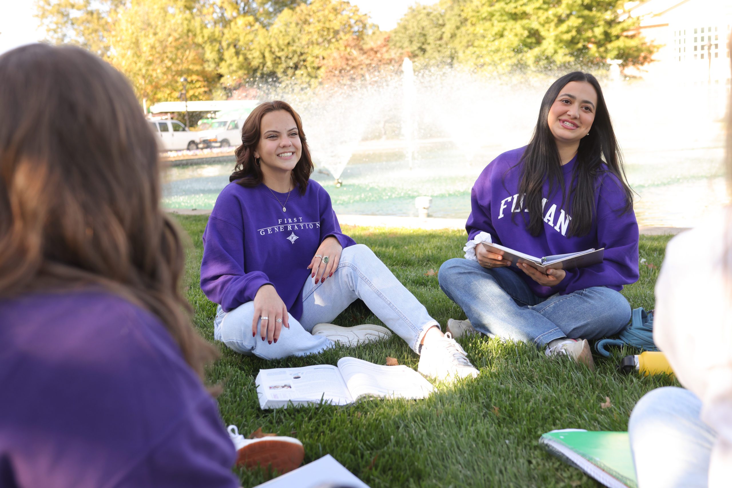 Female students laughing together and sitting in the grass in front of a water fountain on campus