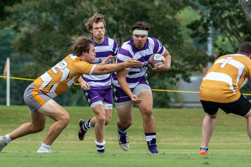 Furman rugby player runs with the ball tucked, a teammate by his side, and a GA Tech player crouching for a tackle.