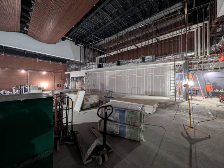 A construction picture shows a wide angle view of the future functional fitness area. The field is littered with active construction materials and the background highlights a drywall enclosure for future weight equipment.