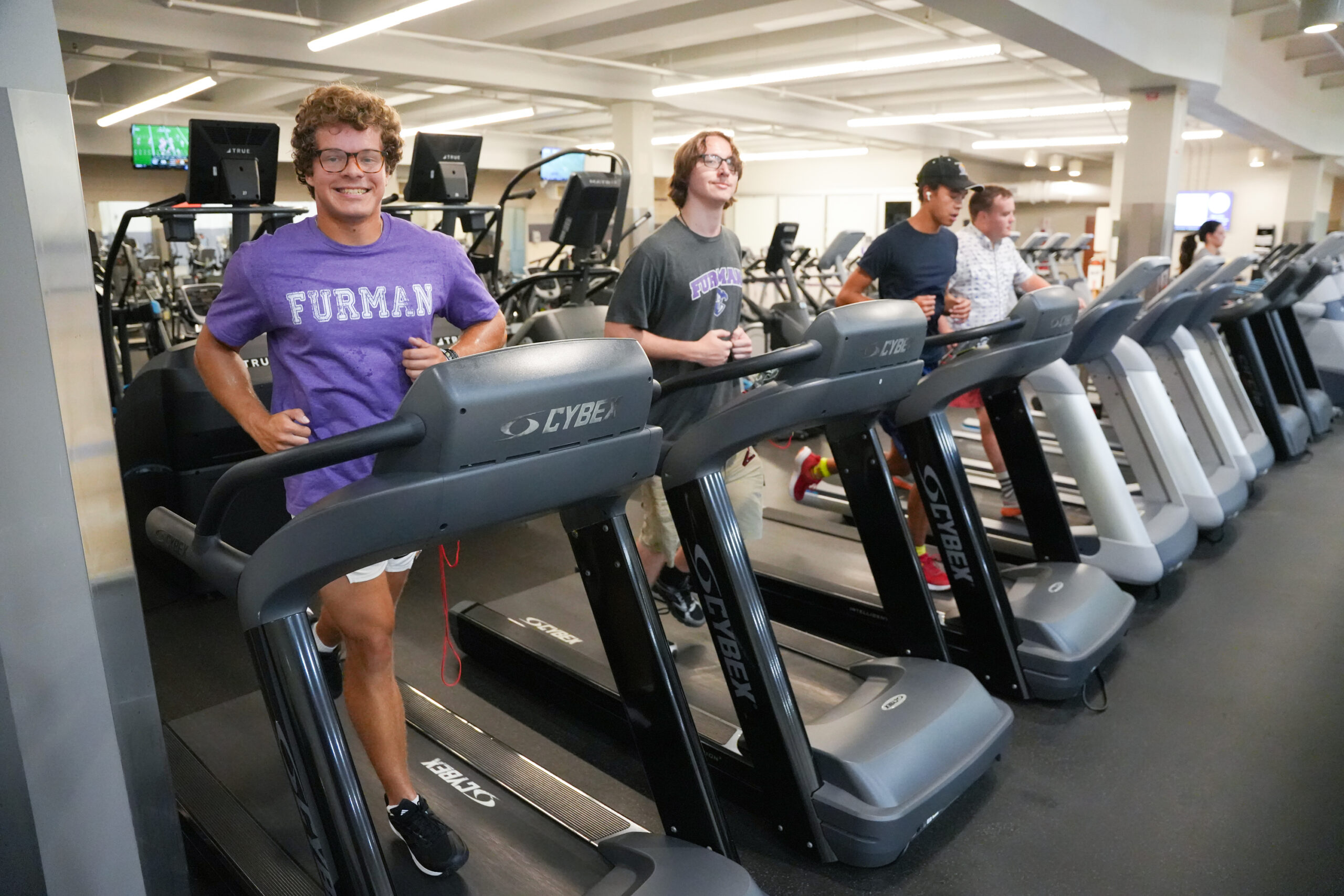 Student smiles broadly while walking on the first treadmill in a line of occupied treadmills.