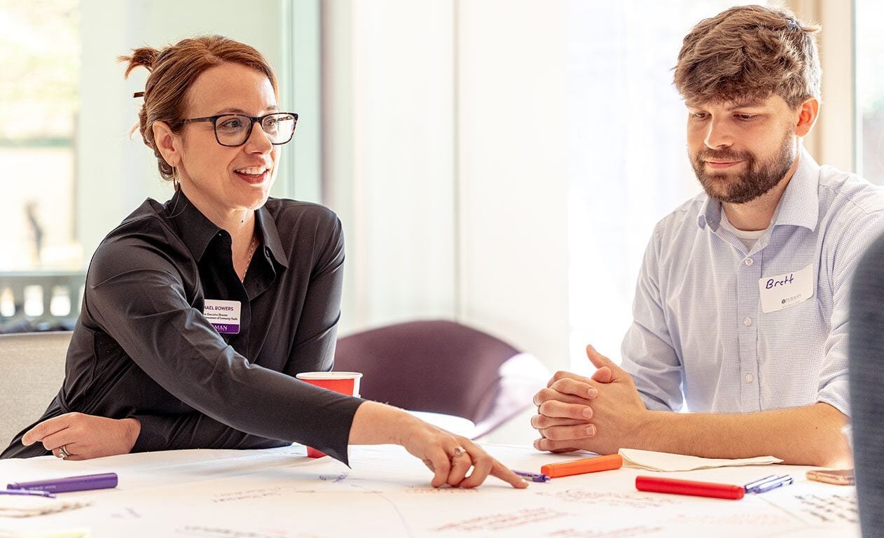 a woman points to a poster board on a round table while speaking to the group