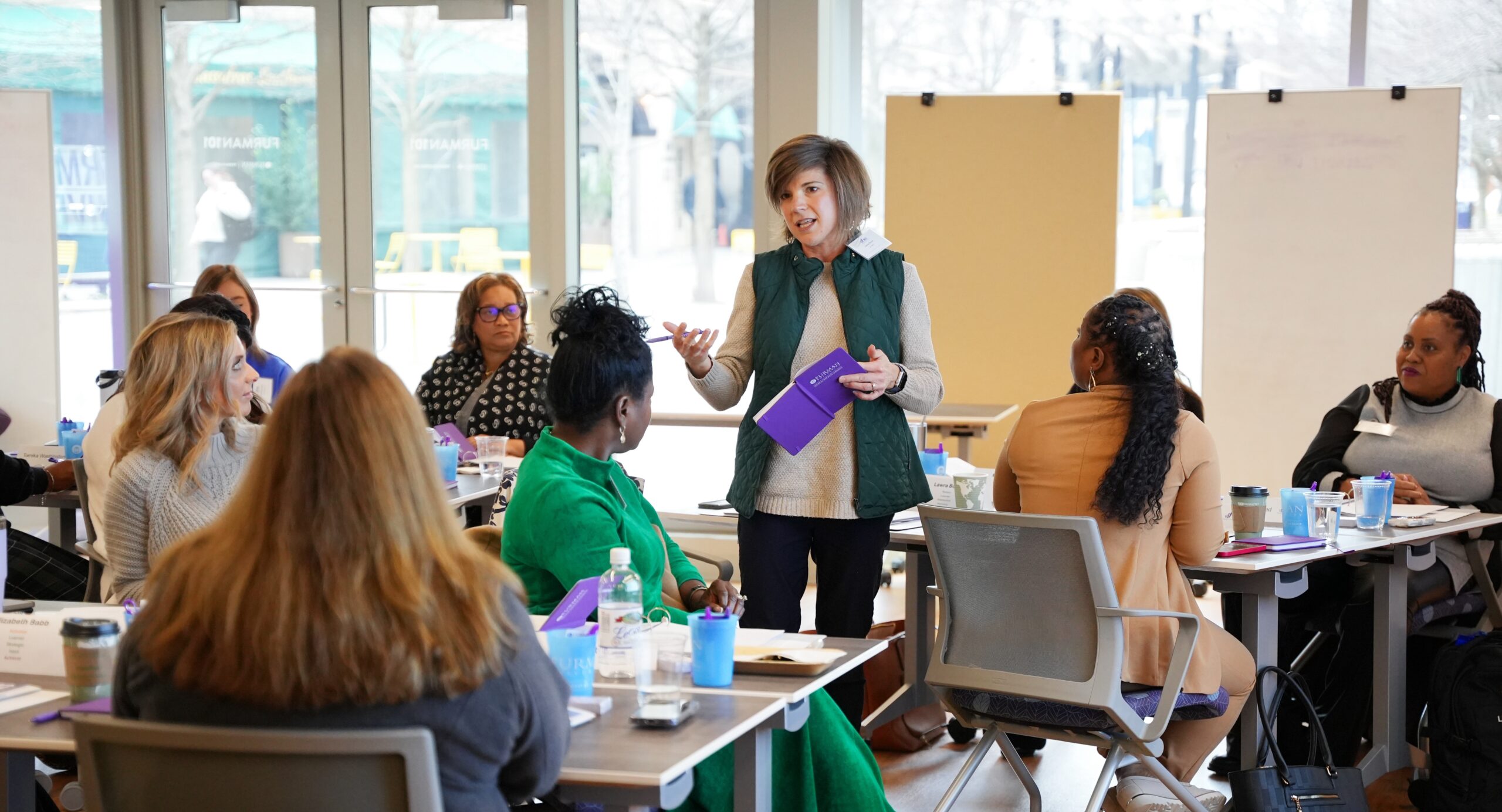 A group of diverse women are sitting listening to one standing woman providing explanation