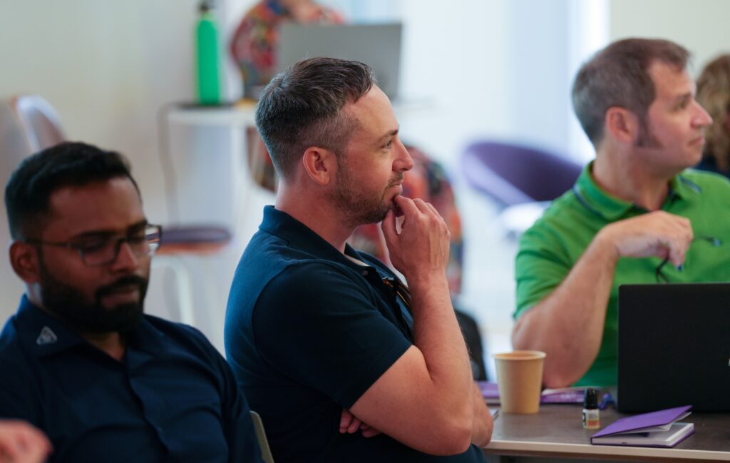 three men are seated looking at a presenter off screen. the camera is focused at the one in the middle, who is listening with a thoughtful expression.