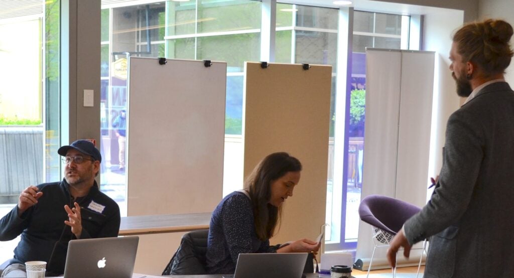 A man actively discussing with a woman seated at a table with her laptop in front of her. A different man seated next to her is in conversation with someone off the screen.