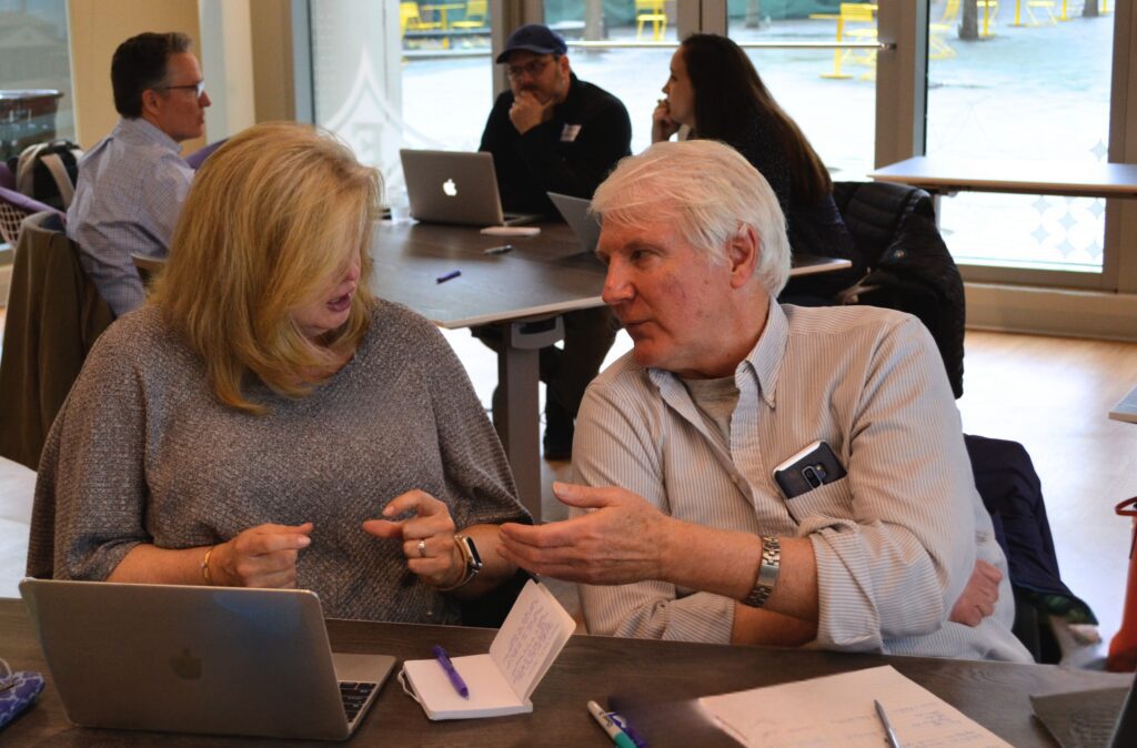 A man and woman are discussing in front of a laptop screen, others can be seen sitting behind them doing similar