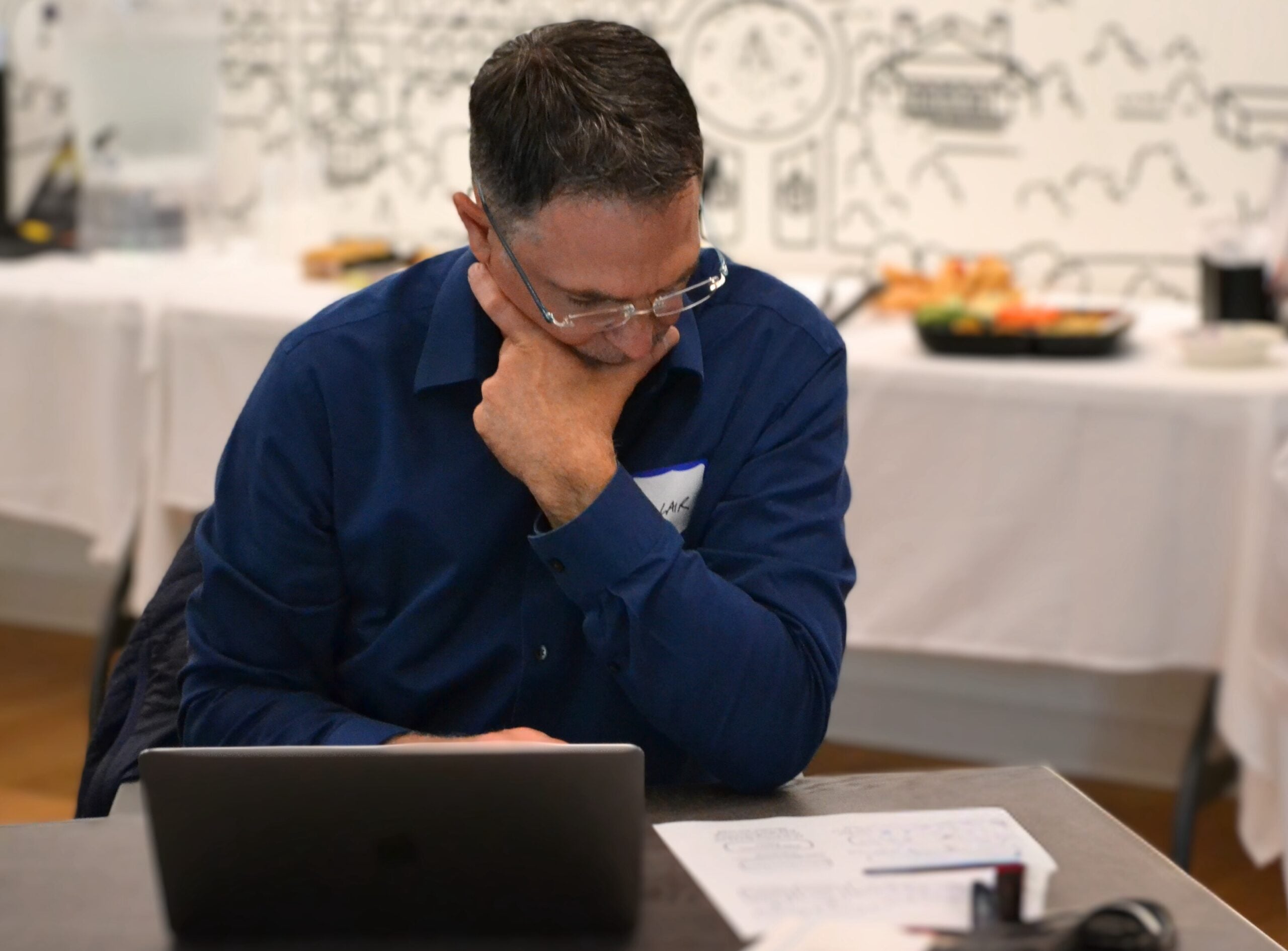a lone is sitting at a table, thoughtfully staring down at his laptop and working area.