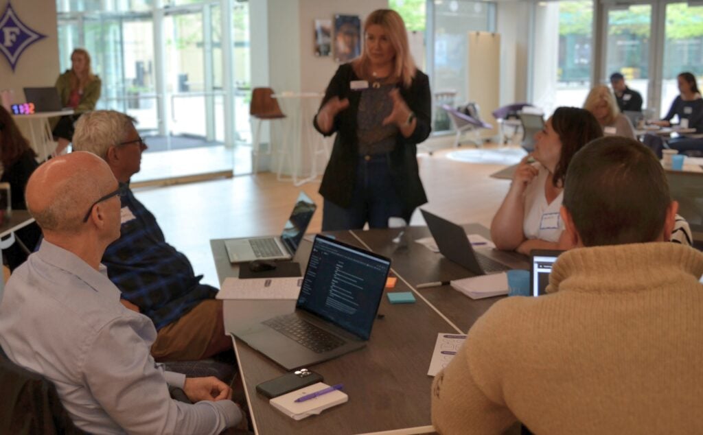 a group of different individuals are seated at a table with laptops open, looking at one woman standing and gesturing with her hands.