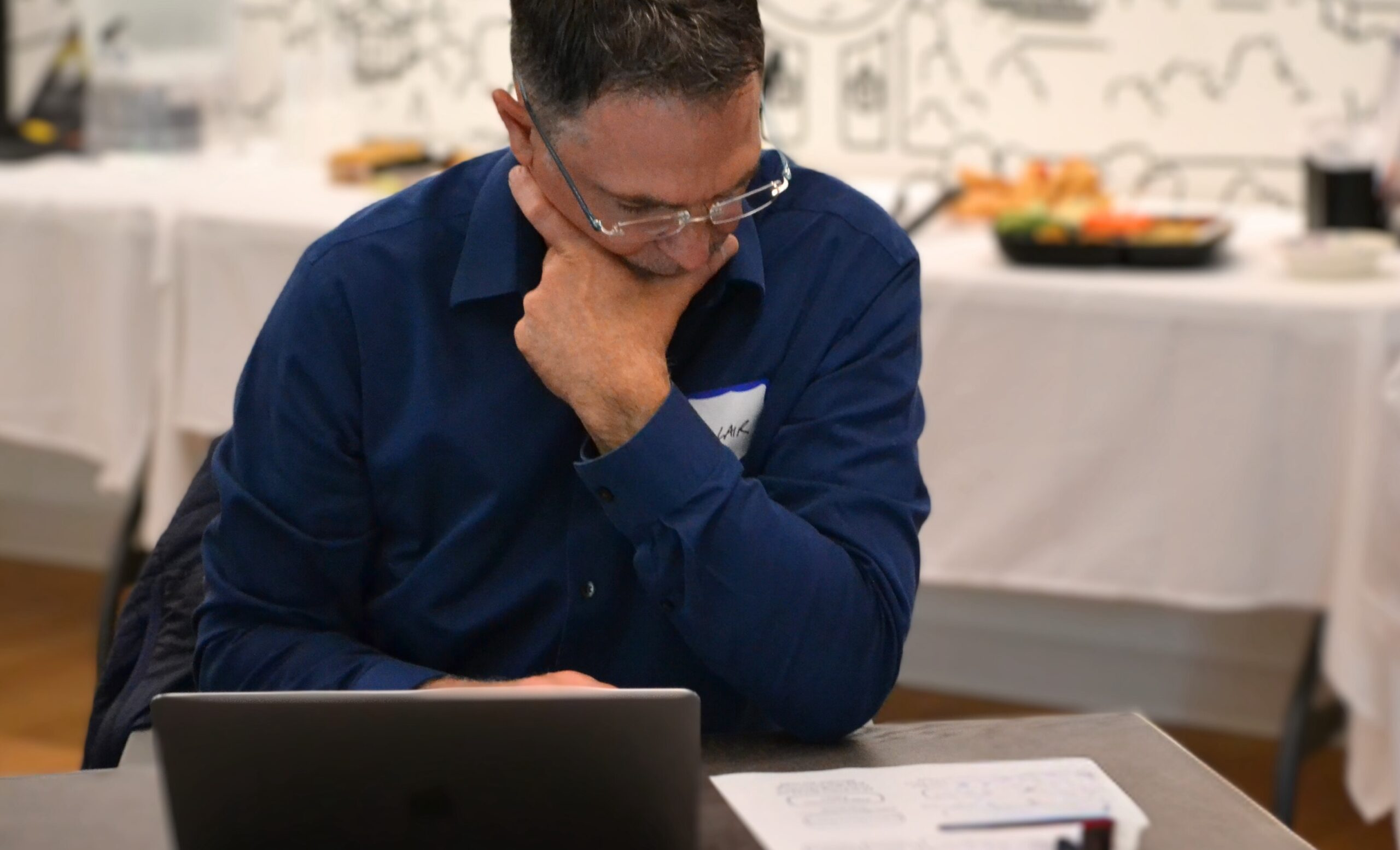 a lone is sitting at a table, thoughtfully staring down at his laptop and working area.