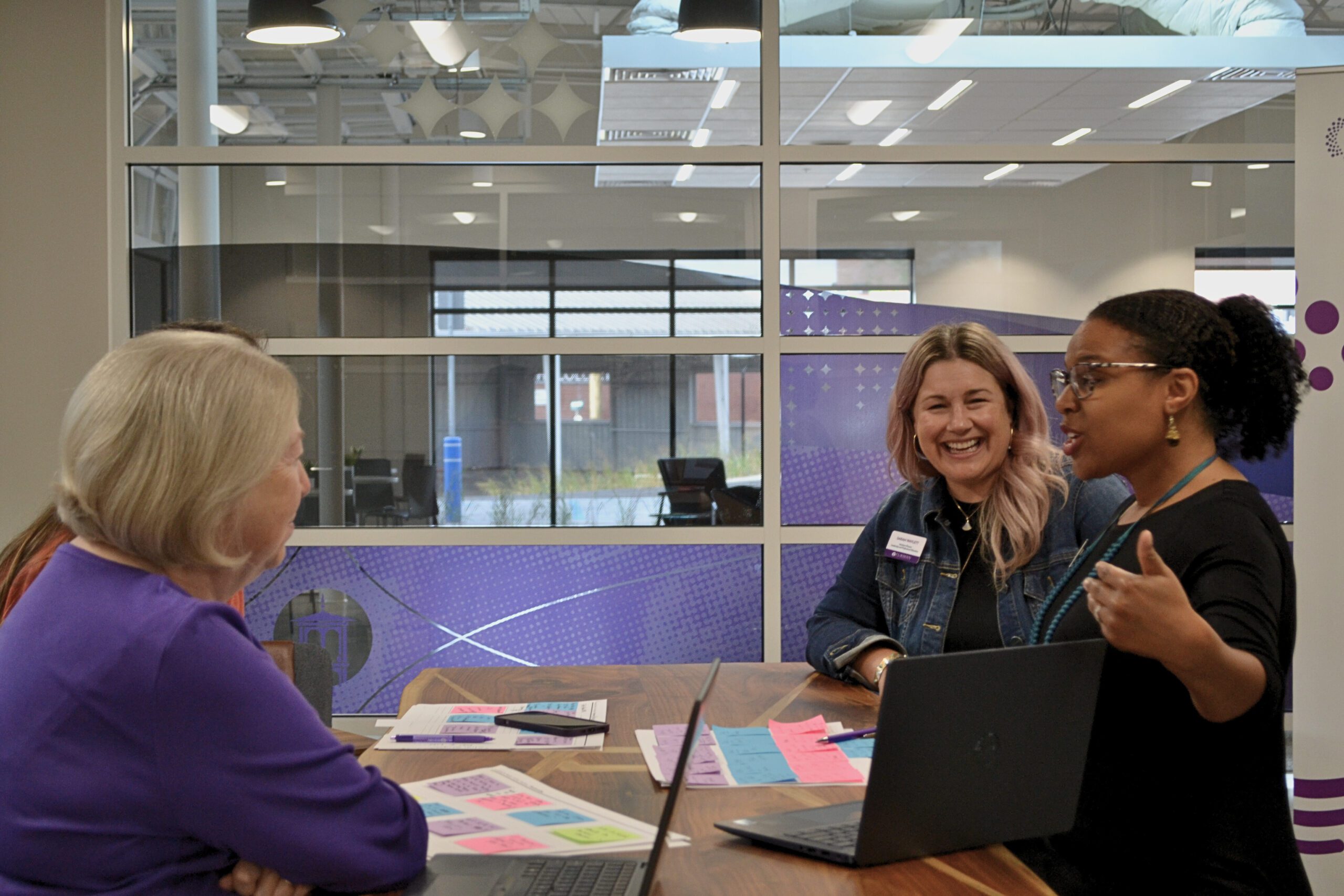 A group of diverse women are sitting at a table with signs of collaborative work with post-its and laptops, listening to an African American woman speak.