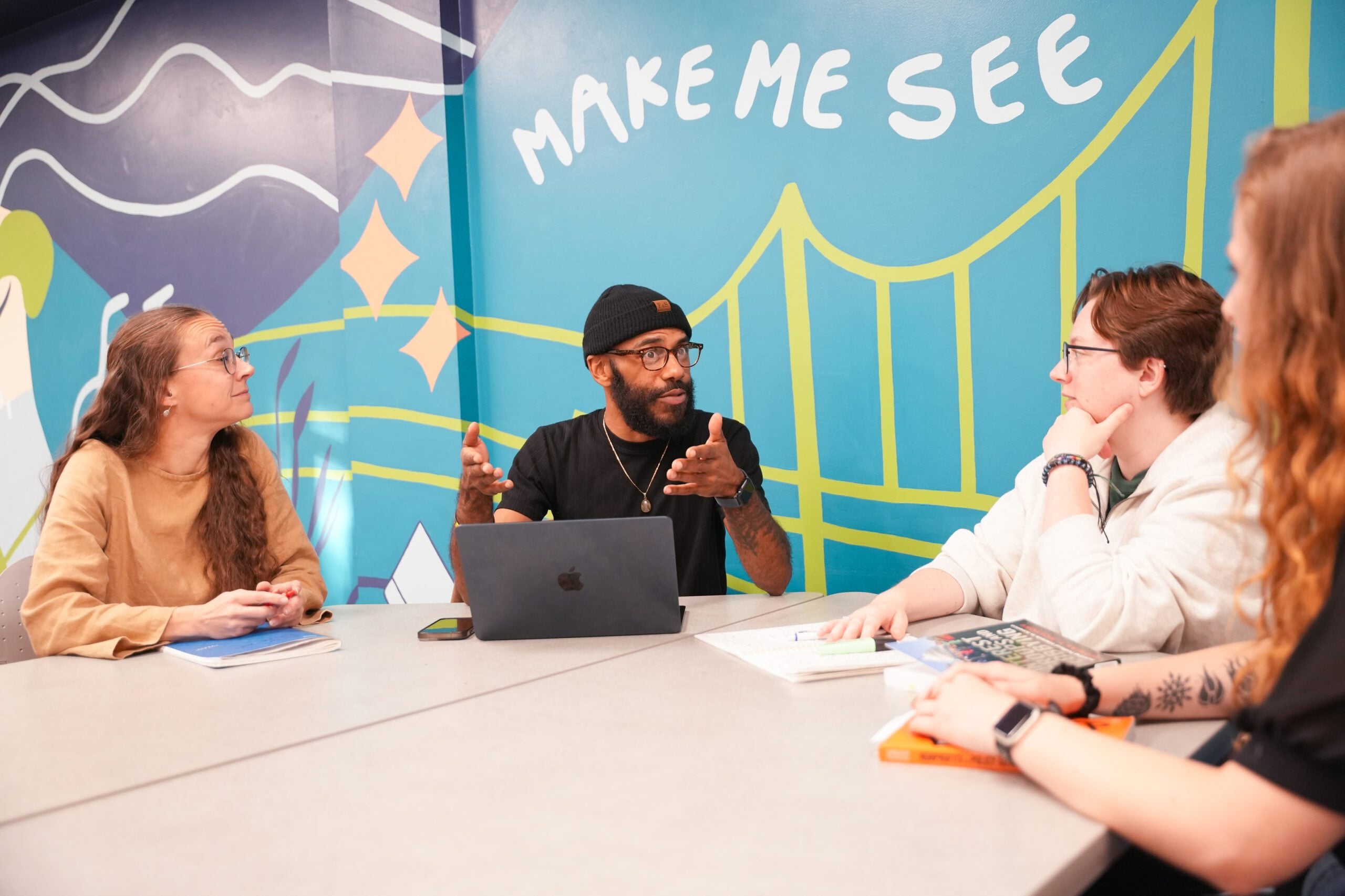 Four students sit around a conference table during a discussion. One participant gestures while speaking from behind a laptop as others listen and take notes.