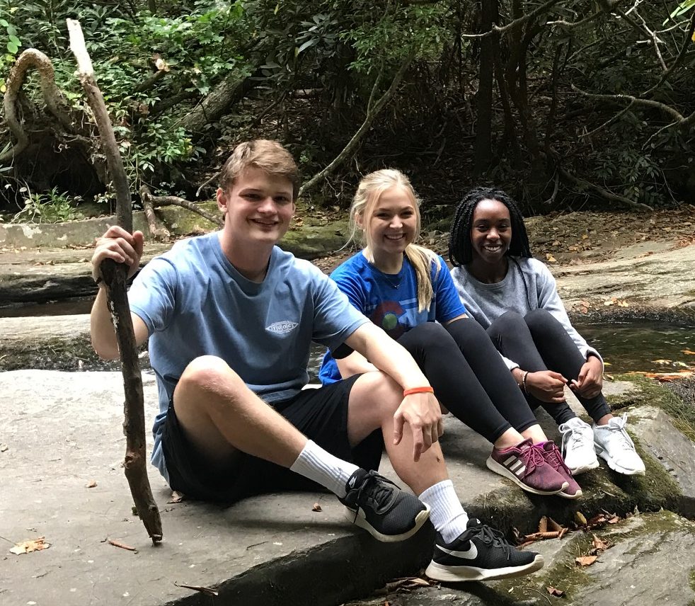 Three students sitting on a rock beside a stream in Upstate park.