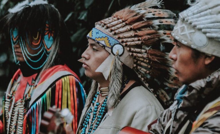 Three Native American individuals in traditional ceremonial clothing.