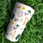 An insulated tumbler with illustrated icons rests on bright green grass, shown from above.