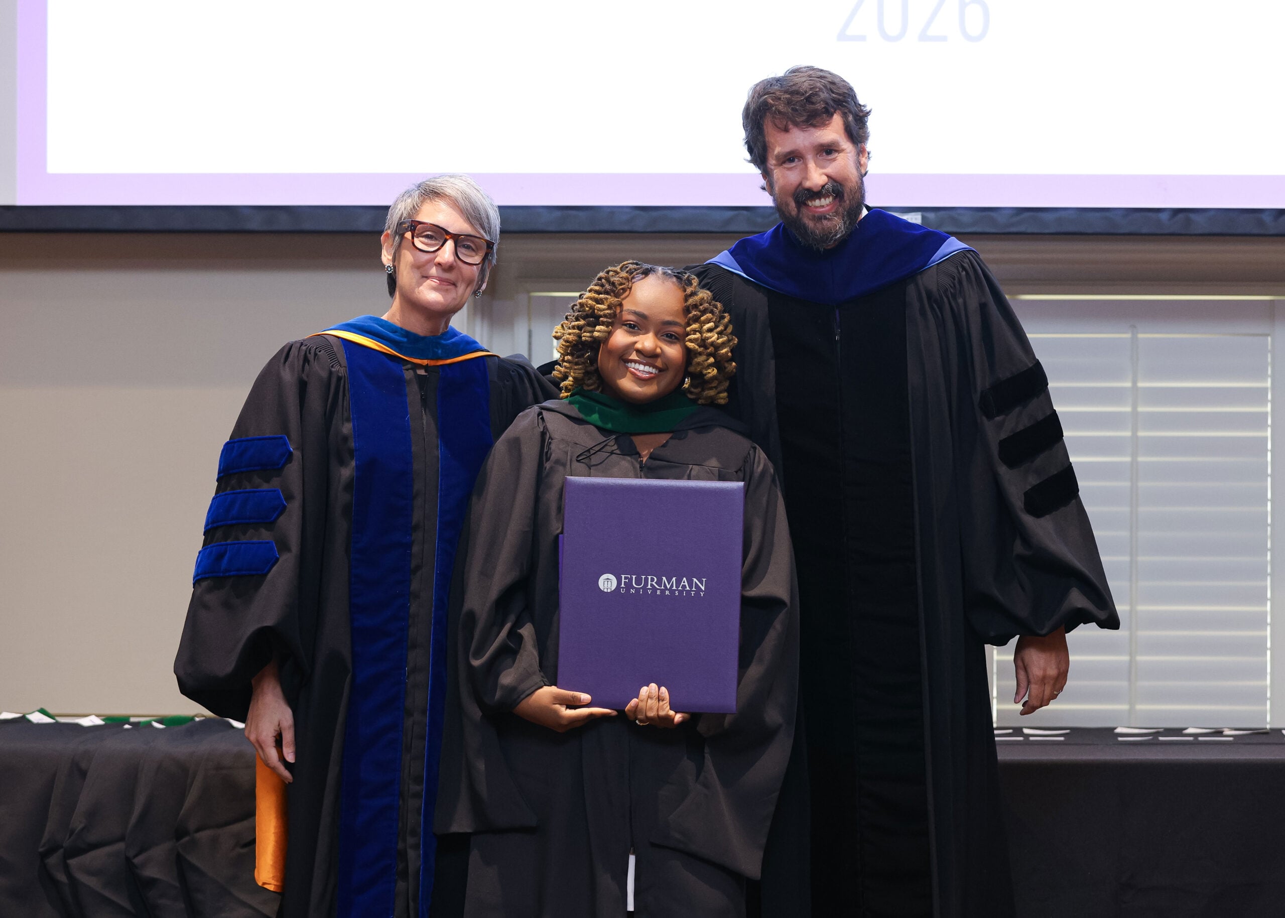 Three people stand together in graduation attire, one holding a degree folder,