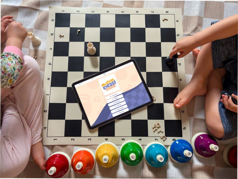Overhead view of two children sitting around a large chessboard mat with chess pieces and musical-note-shaped game pieces scattered across the board. A tablet displaying the Musical Chess learning app rests in the center of the board. Along the bottom edge, a row of brightly colored handbells arranged in rainbow order, labeled with musical notes, sits on a checkered tablecloth. One child holds a black chess piece while interacting with the game, which combines music learning and chess concepts.