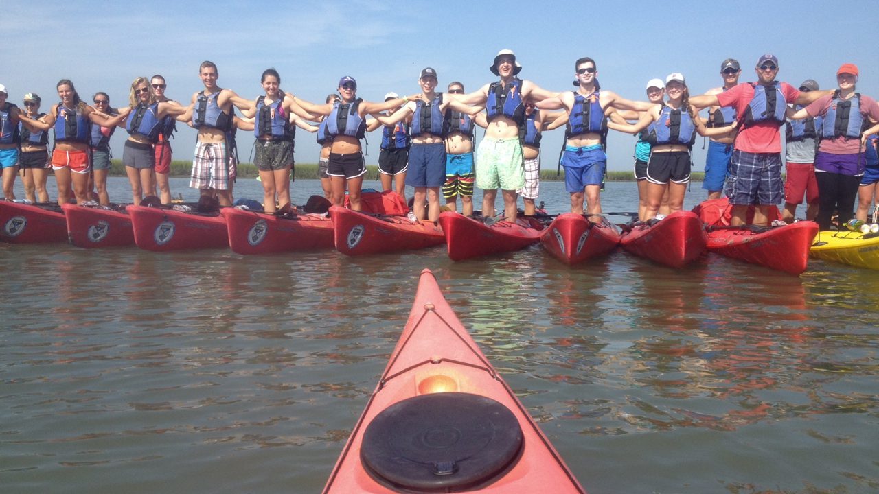 Students standing on a row of kayaks, holding onto each other's shoulders