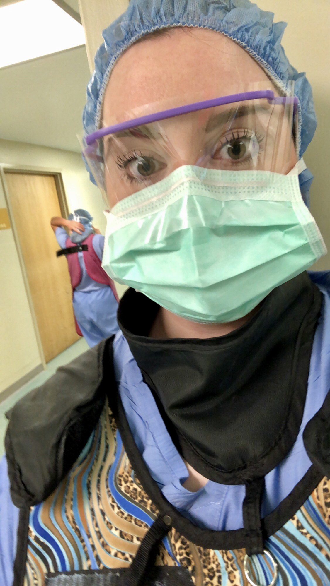 Selfie of a woman in scrubs and personal protective equipment in a hospital corridor.