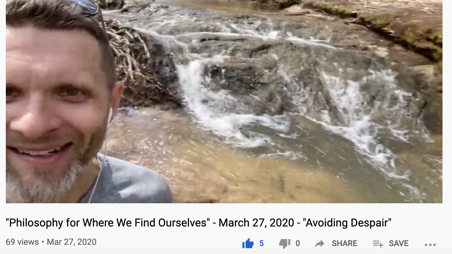 A man with a goatee shoots a video selfie in front of a large creek with rapids.