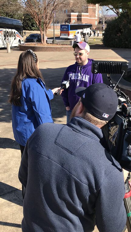 Outside, a man in purple is interviewed by a TV reporter and a videographer.