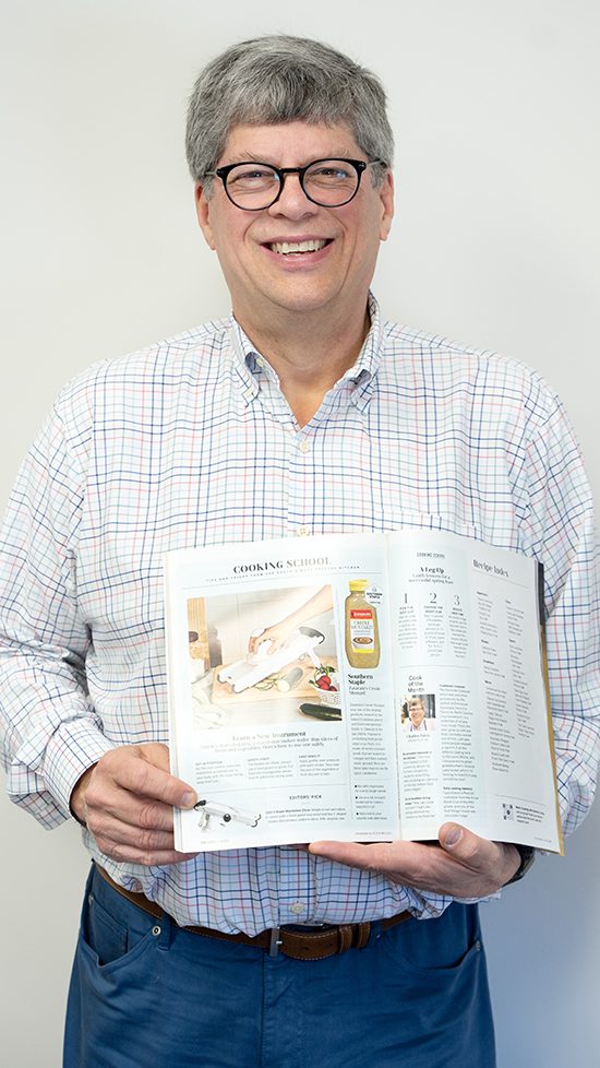 A man dressed in jeans and a long sleeve shirt holds a magazine and smiles.