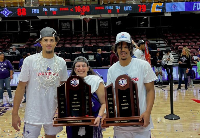 Two Black basketball players flank white woman holding trophy
