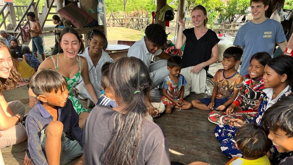 Furman students and Cambodian children sit in a circle smiling and laughing.