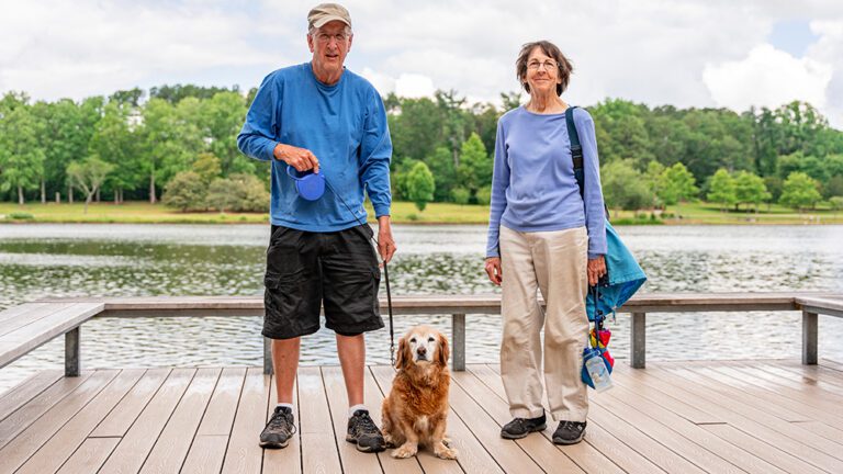 Photo of a man, a small dog, and a woman, standing in front of Furman Lake. 