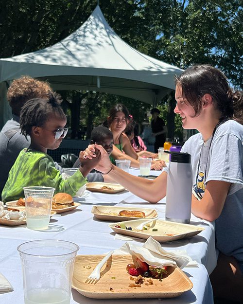 A young Black girl is seemingly about to arm-wrestle with a young white woman while seated at an outdoor picnic table covered in dishes and a tablecloth.
