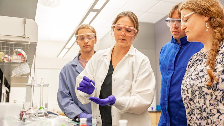 Three students and a professor wearing protective goggles look at a sample in a chemistry lab.