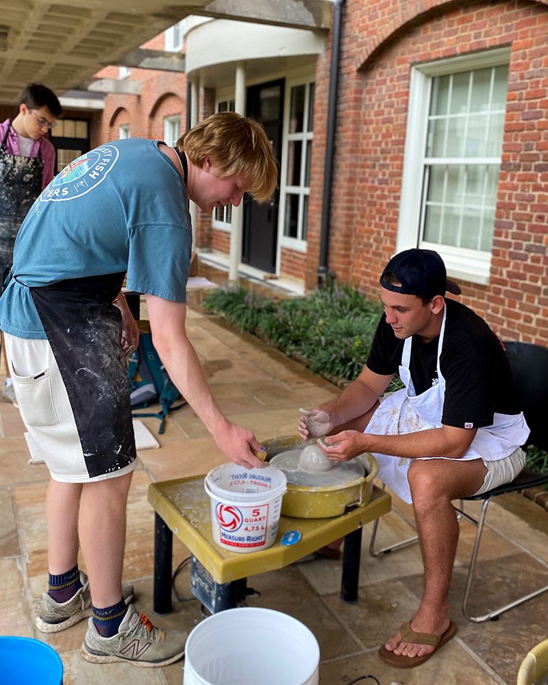 One male student bends to help another male student seated at a pottery wheel.