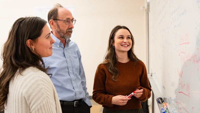 Two female students and a male professor discuss what's on a white board.