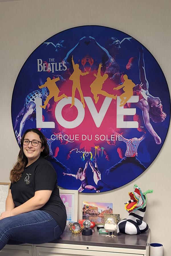 Woman sits and smiles in front of taken her math skills and love of the entertainment world to one of the most well-known, premiere circuses — Cirque du Soleil poster.