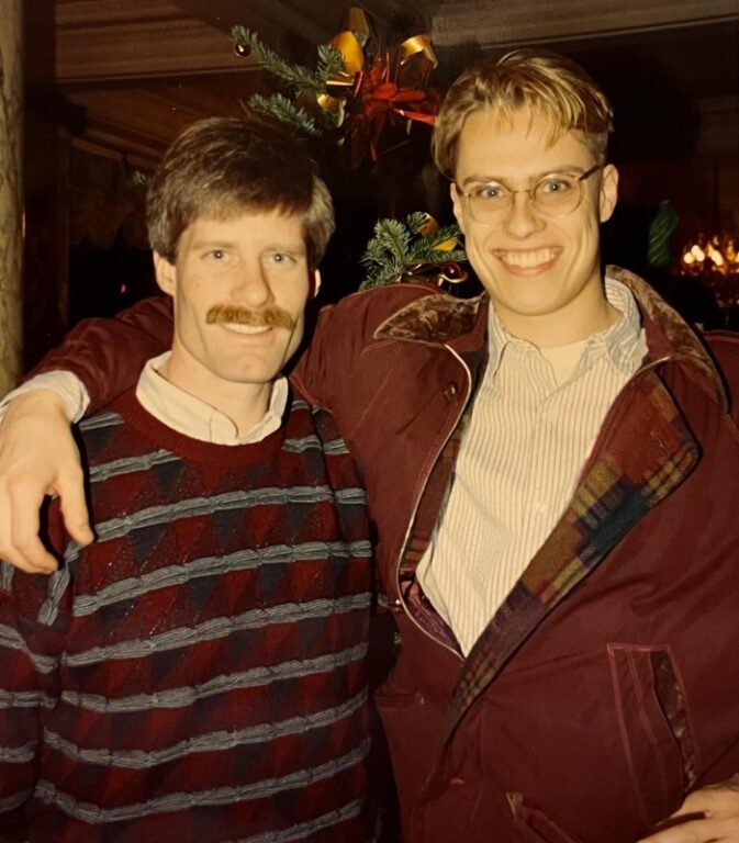 Old photo (1994) of two men smiling at the camera. One wears a mustache, the other, glasses.