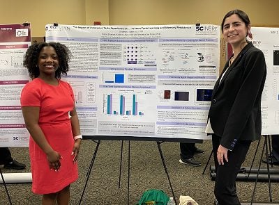 a Black woman in coral dress and a white woman in dark suit stand at poster