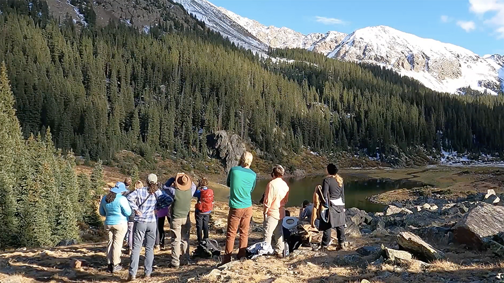 Screen shot of video, students with back to the camera standing at a river at the foot of beautiful mountains.