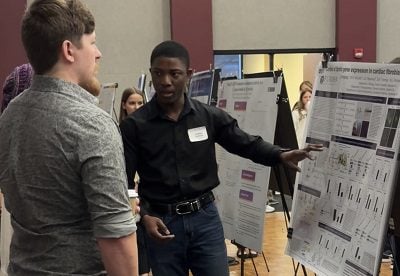a white man speaks with a Black man who is pointing to poster