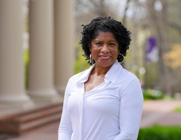 Portrait of a woman standing outside. Large columns of a building to the left. She is Black, wearing a white blouse and silver hoop earrings.