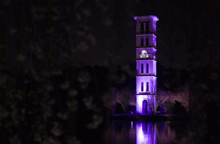The Furman Bell Tower at night with purple lights and a face Taco Bell Tower logo.