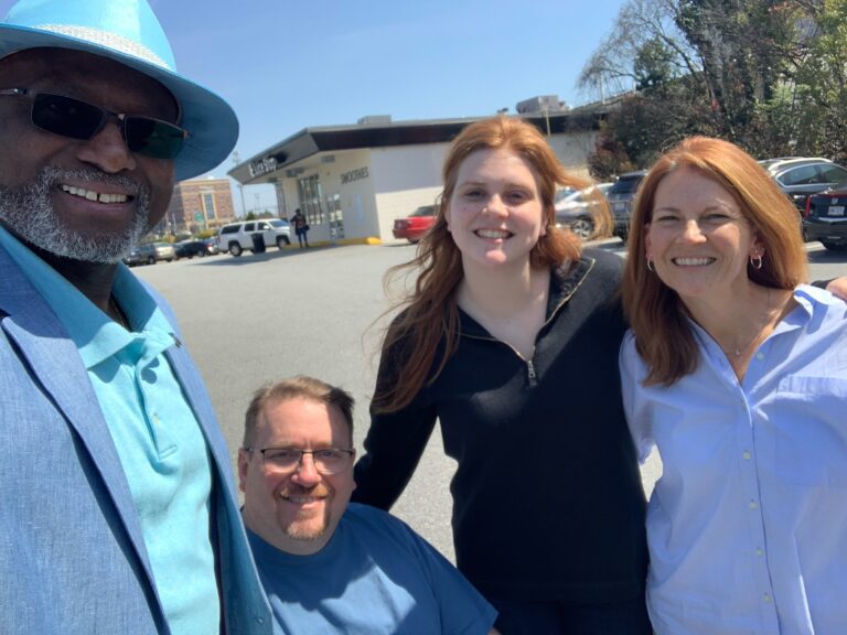 Four people pose for a selfie in a parking lot on a sunny day. A Black man in a suit and hat, a white man in a wheelchair, a young woman with red hair and a woman with red hair. 