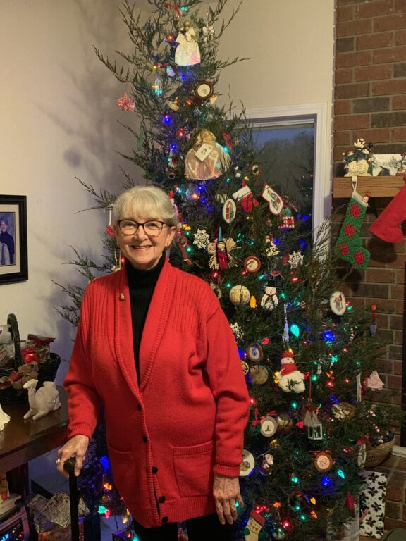 An older woman in a red sweater stands in front of a fully decorated Christmas tree. 