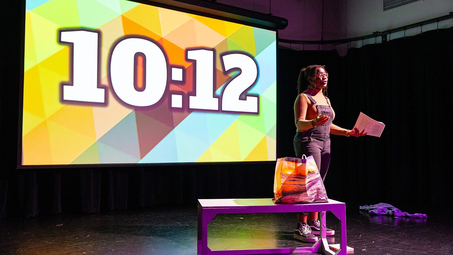 A black woman wearing overalls stands on stage in front of a colorful screen that reads 10:12