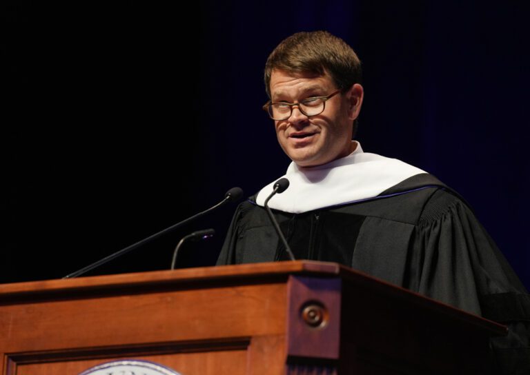 A white man wearing glasses and a graduation robe talks at a lectern