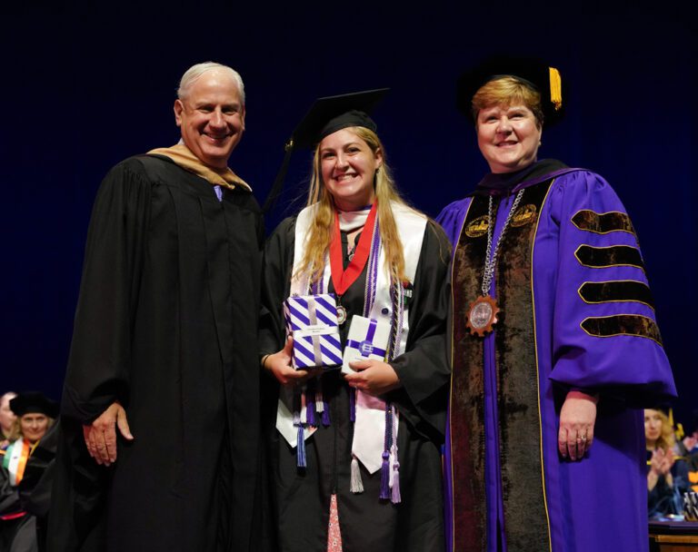 An older white-haired white man, a young woman with long blonde hair and a woman with short blonde hair stand in graduation regalia for a photo. The young woman holds two wrapped packages.