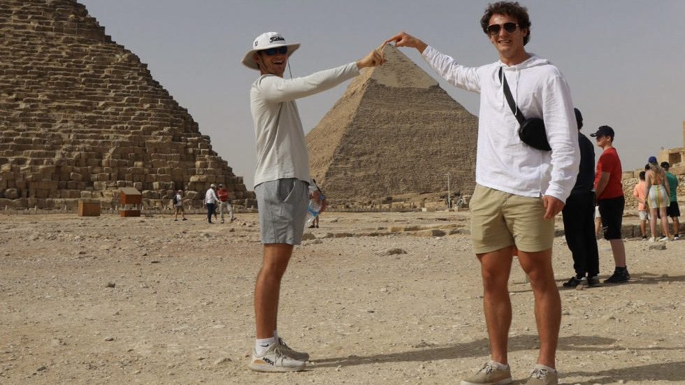 Two young White men pose in the desert with the pyramids behind them. They're pointing to the top of a pyramid.