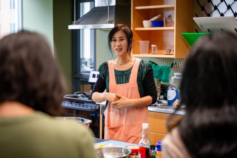 A Korean woman stands in front of a table of ingredients wearing a peach -colored apron.