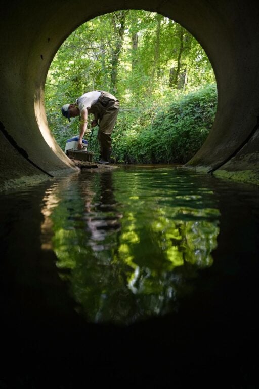 A man is seen at the far end of a large culvert where water reflects the light.